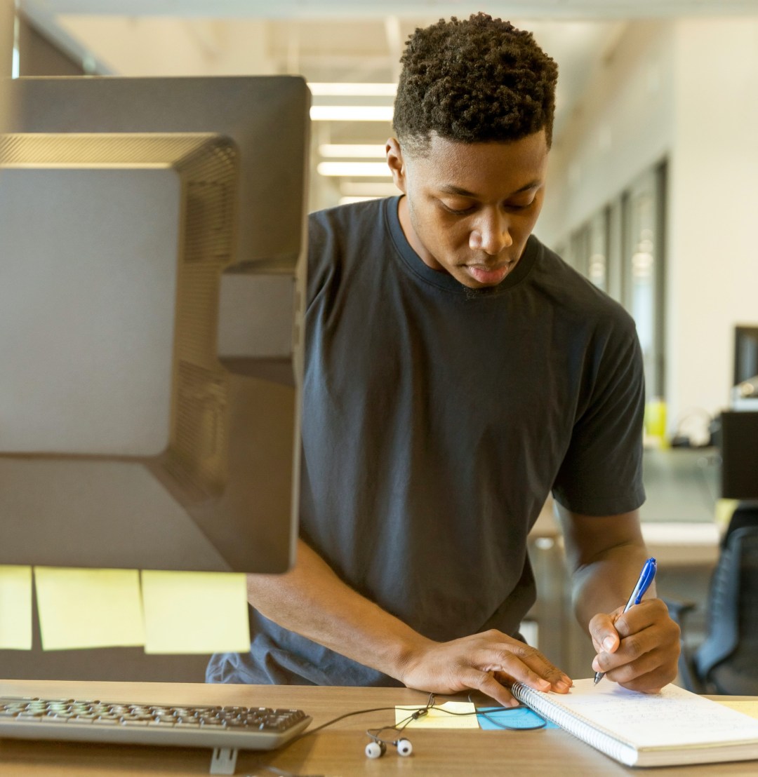 A young man stands at a desk with a computer and writes on a notepad.